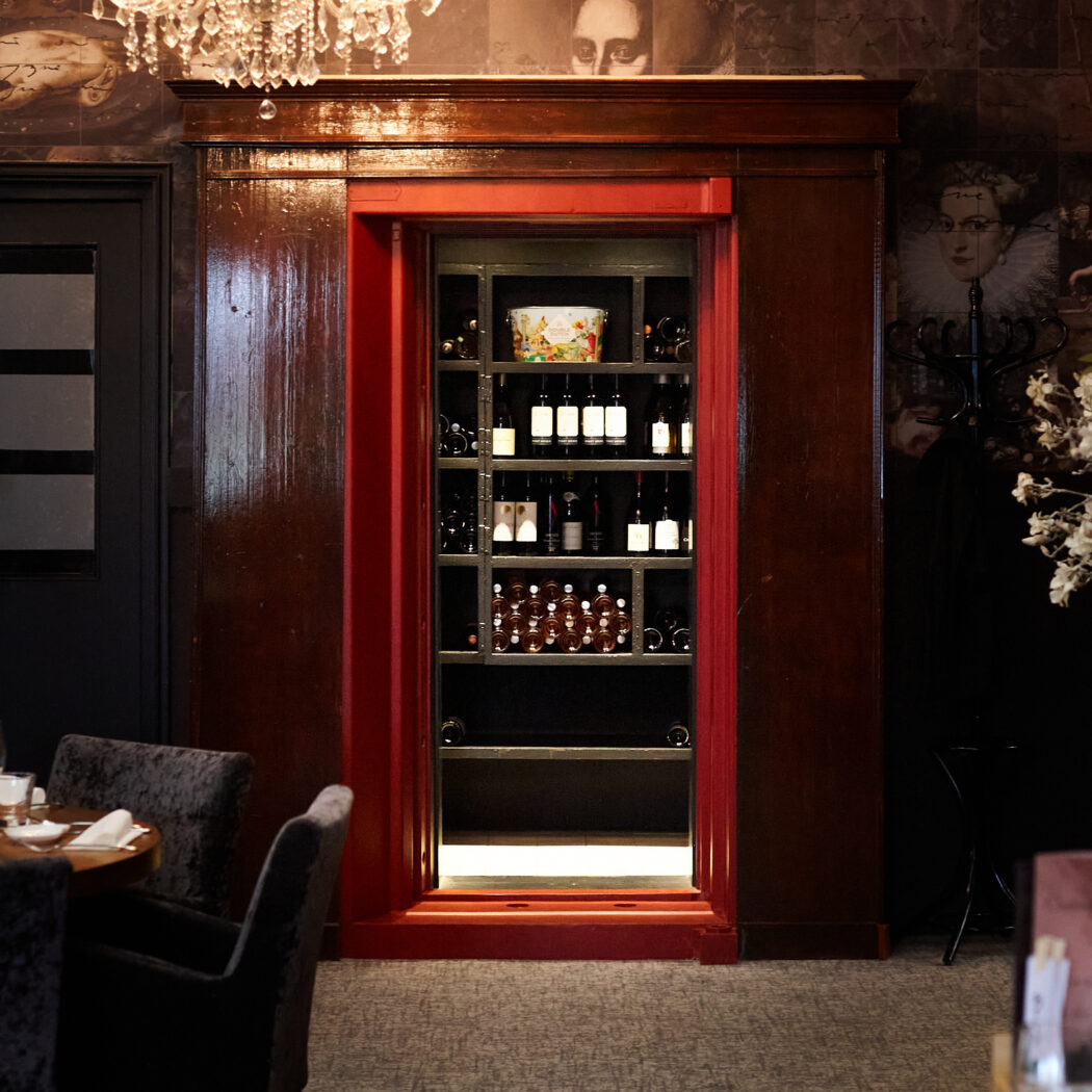 Ingebouwde, verlichte wijnkast met rode omlijsting, flessen wijn, kroonluchter en klassiek behang in restaurant.