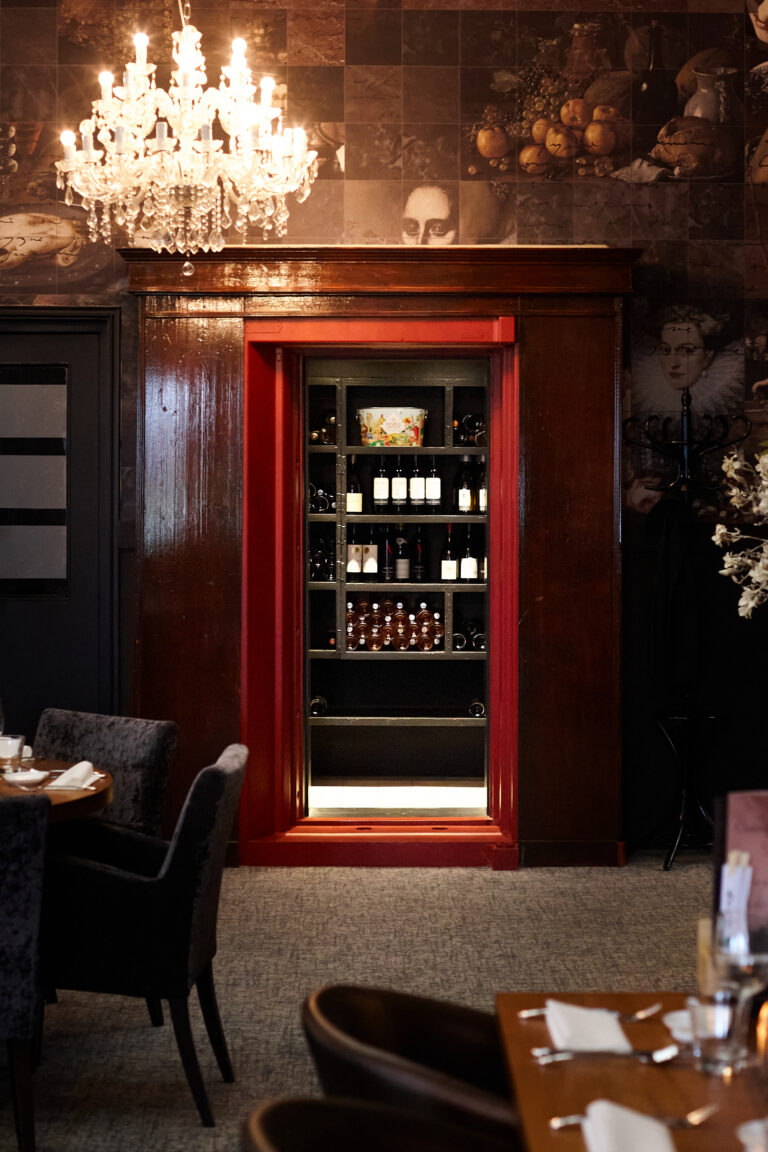 Ingebouwde, verlichte wijnkast met rode omlijsting, flessen wijn, kroonluchter en klassiek behang in restaurant.
