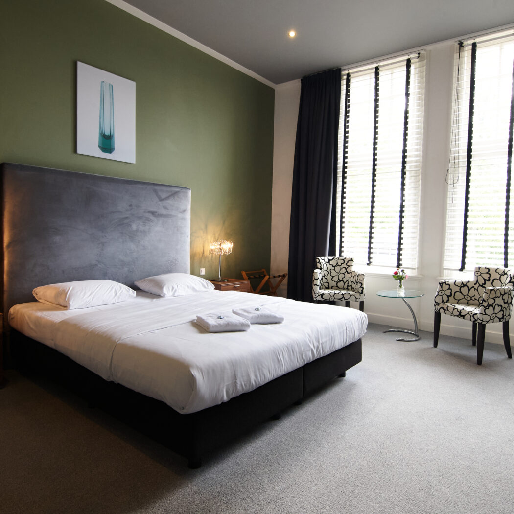 Moderne hotelkamer met comfortabel tweepersoonsbed, groene muur en een zithoek bij de ramen.