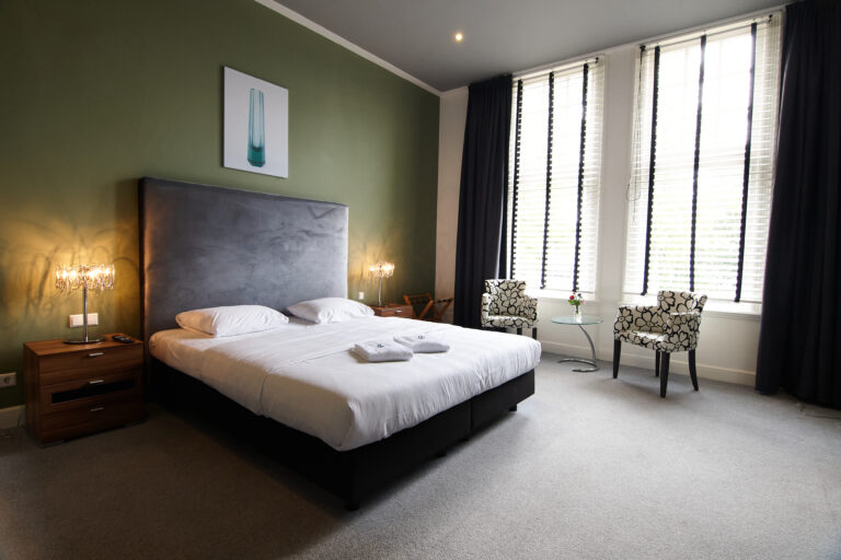 Moderne hotelkamer met comfortabel tweepersoonsbed, groene muur en een zithoek bij de ramen.
