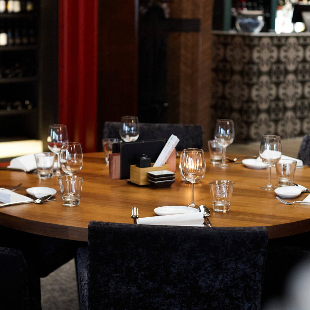 Ronde houten eettafel met zwarte fluwelen stoelen, gedekte plaatsen en een kaars in een sfeervol restaurant.