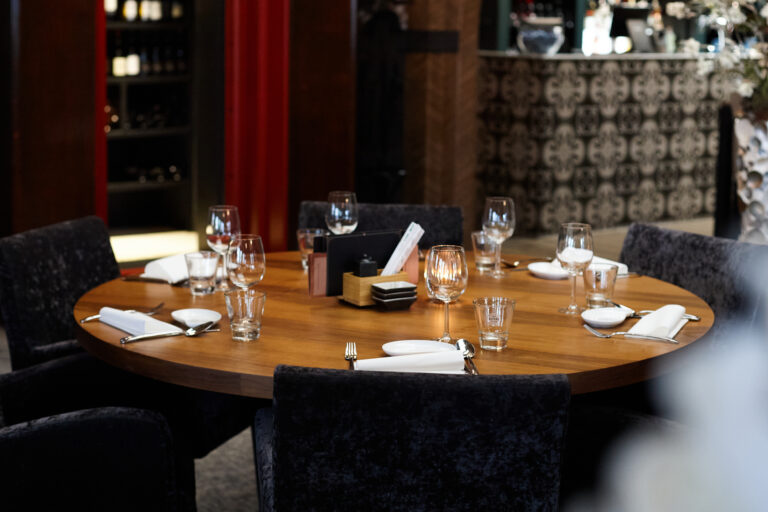 Ronde houten eettafel met zwarte fluwelen stoelen, gedekte plaatsen en een kaars in een sfeervol restaurant.