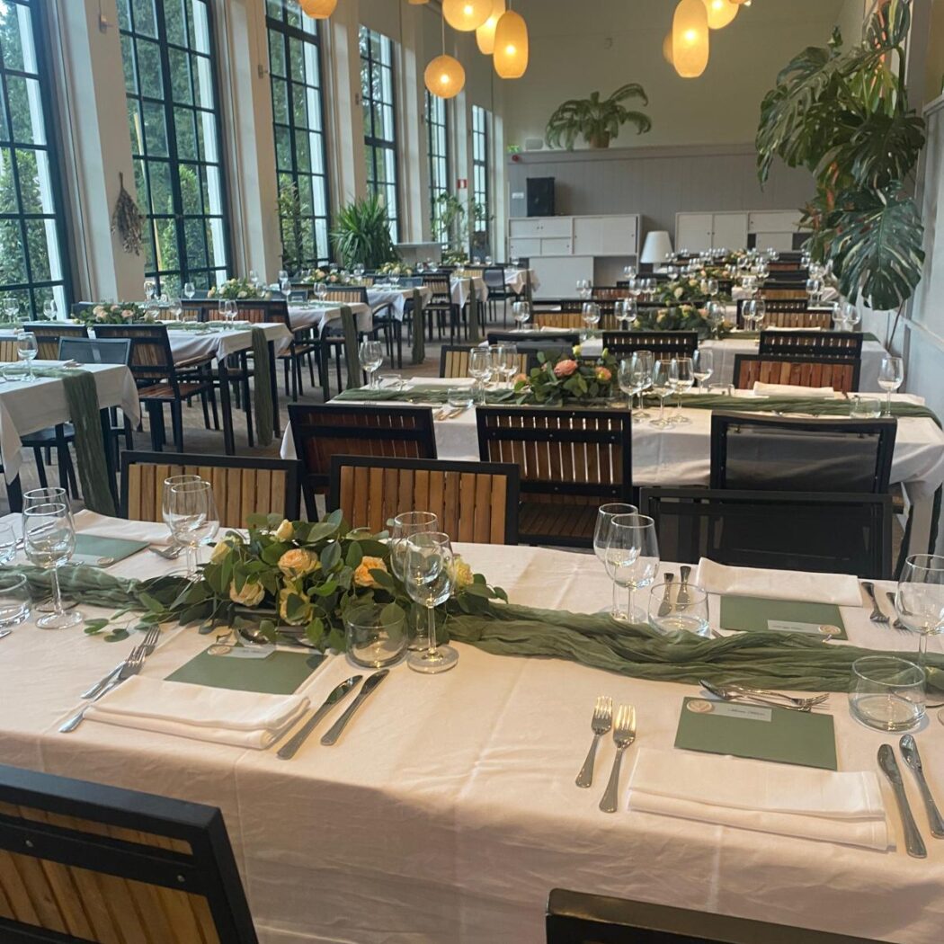 Elegante feestzaal met lange gedekte tafels, groene lopers, rozen, hanglampen en grote ramen.