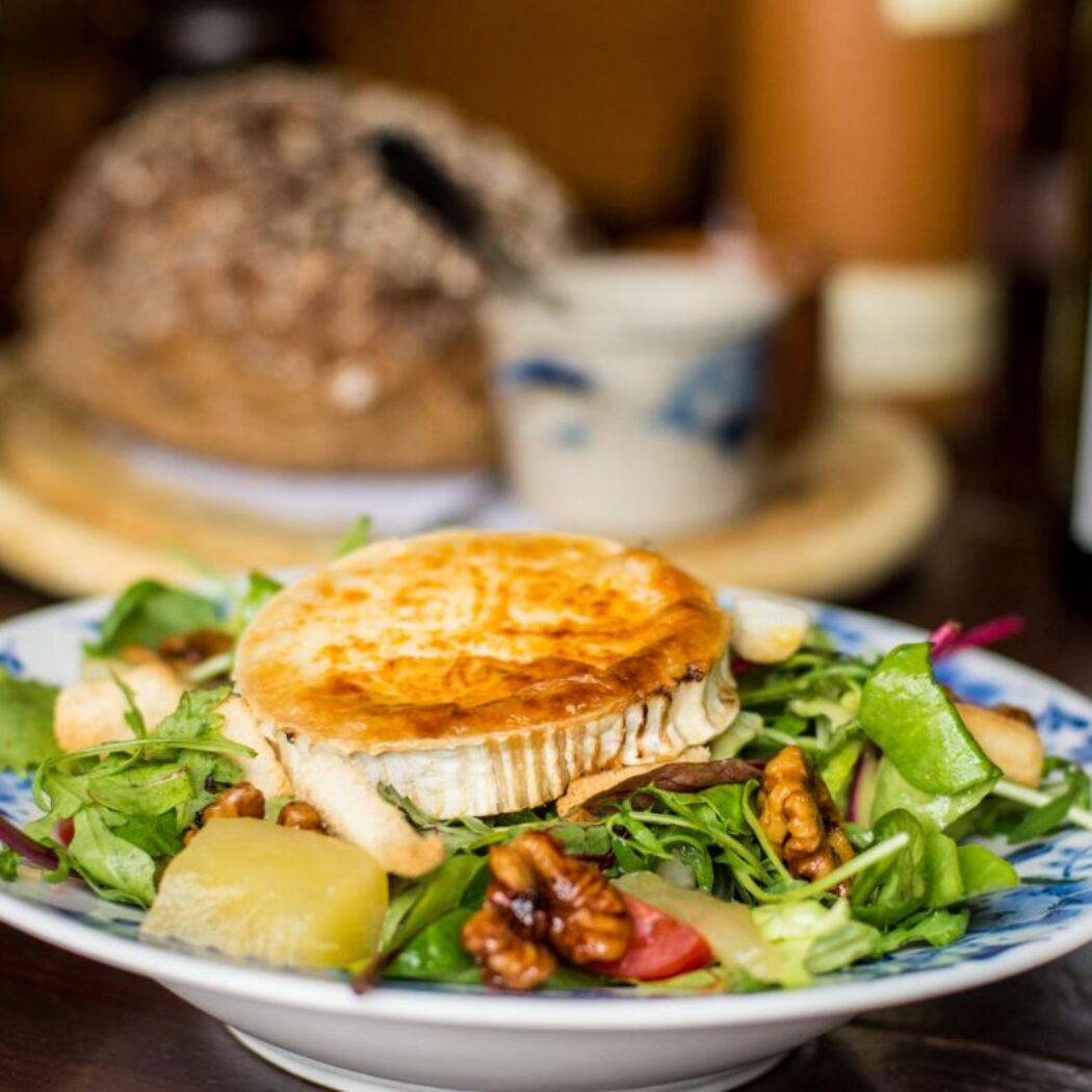 Een bord met verse groene salade, gegrilde geitenkaas, walnoten en peer, met wijn op de achtergrond.
