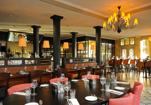 Elegant restaurant met lange ovale tafel, leren zitbanken, een bar en opvallende donkere zuilen en kroonluchter.