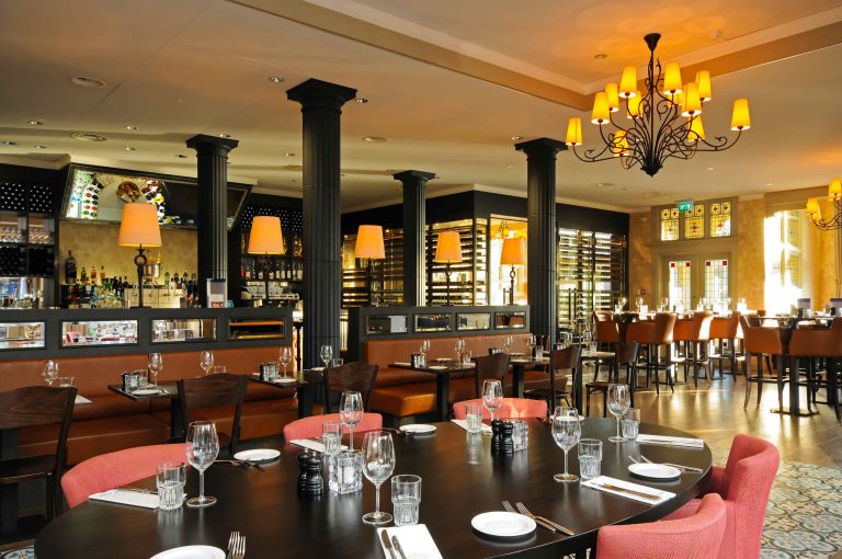 Elegant restaurant met lange ovale tafel, leren zitbanken, een bar en opvallende donkere zuilen en kroonluchter.