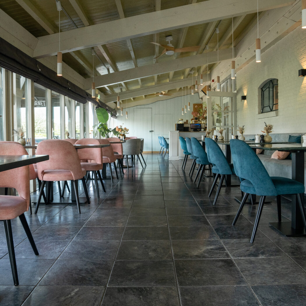 Modern restaurant interieur met roze en petrolblauwe stoelen, donkere vloertegels en grote ramen waar CORNET zichtbaar is.