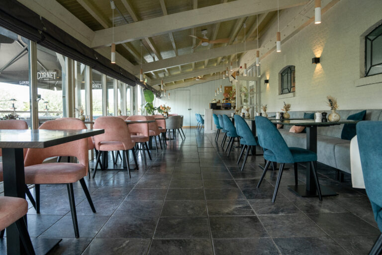 Modern restaurant interieur met roze en petrolblauwe stoelen, donkere vloertegels en grote ramen waar CORNET zichtbaar is.