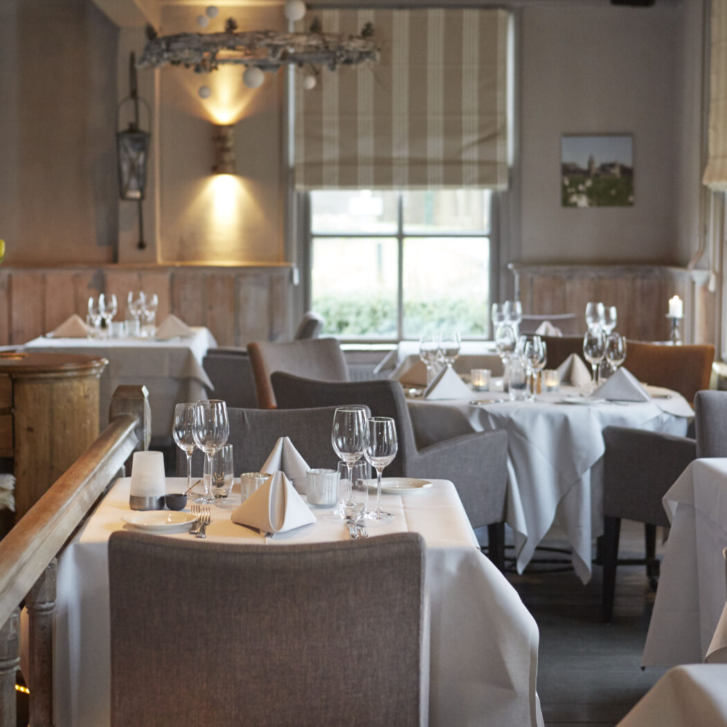 Sfeervol restaurantinterieur met wit gedekte tafels, glazen en servetten, verlicht door zachte wandlampen.