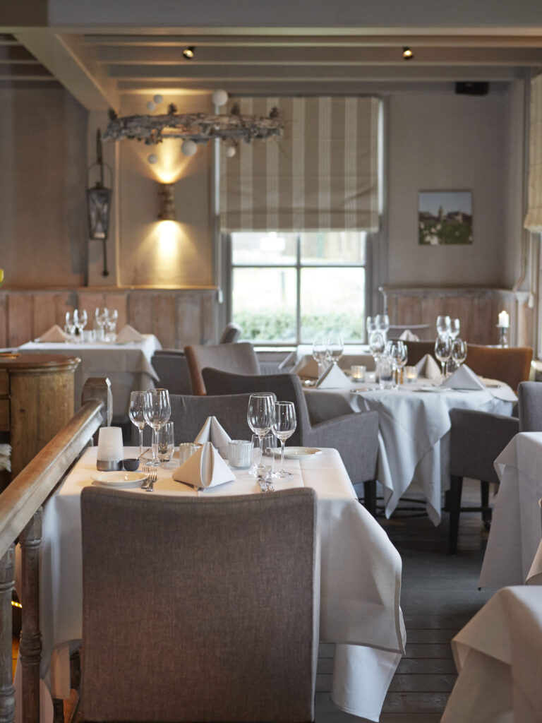 Sfeervol restaurantinterieur met wit gedekte tafels, glazen en servetten, verlicht door zachte wandlampen.