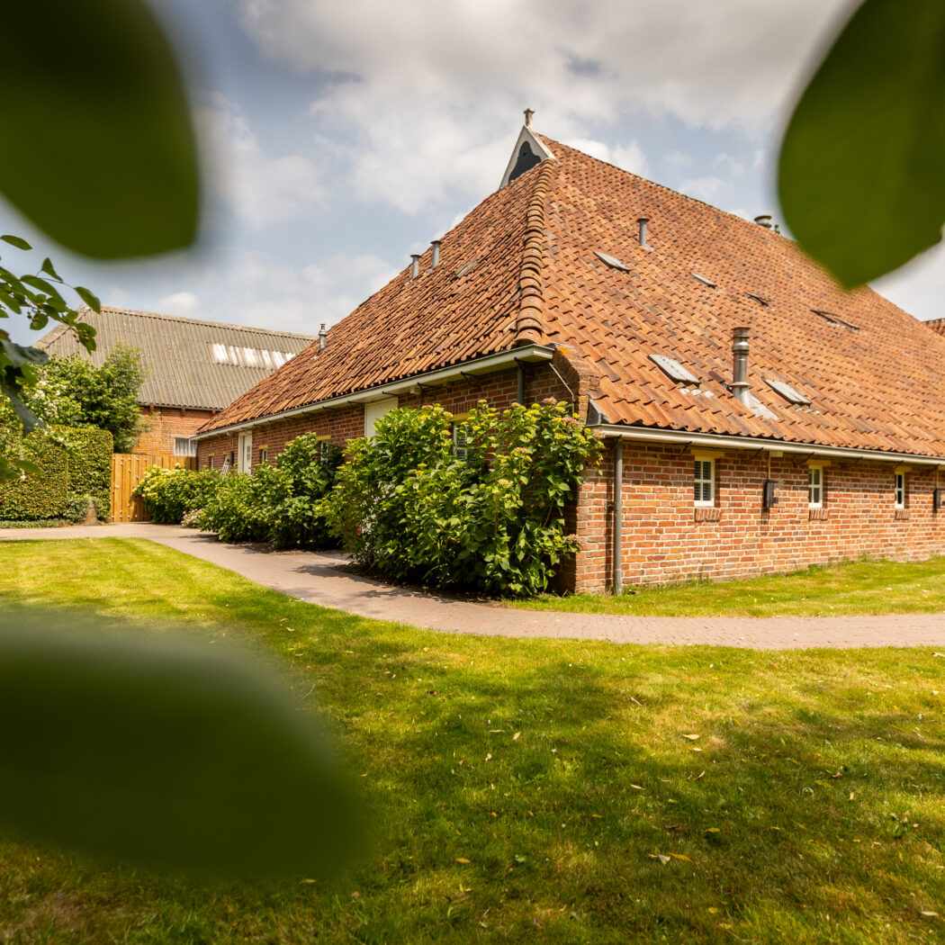 Bakstenen boerderij met steil dak van rode pannen, dakkapellen en groene tuin met kronkelend pad.
