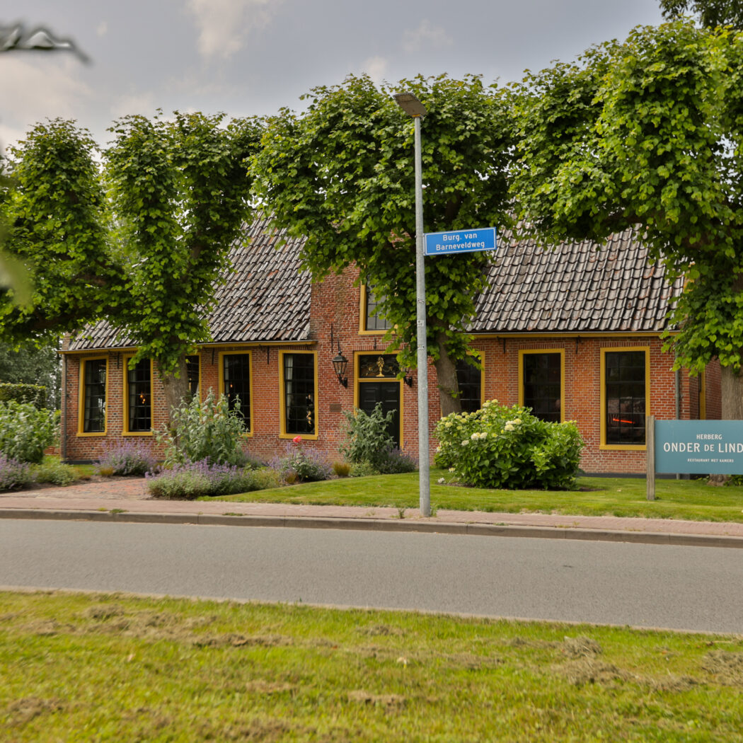 Rood bakstenen gebouw van Herberg Onder De Linden, met gele kozijnen en lindebomen, langs de Borg van Barneveldweg.