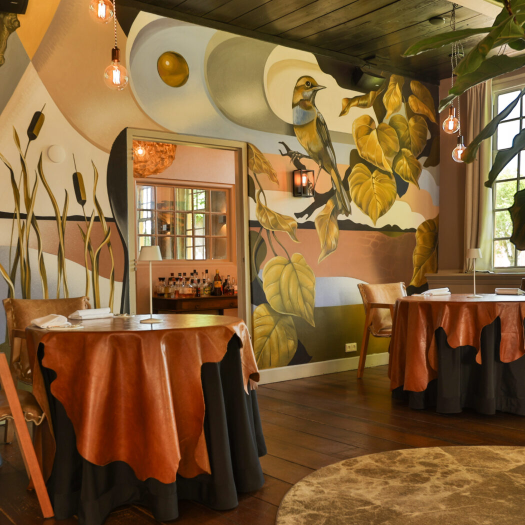 Elegant restaurant interieur met opvallende muurschilderingen van vogels en bomen, en ronde tafels met bruine leren overlays.