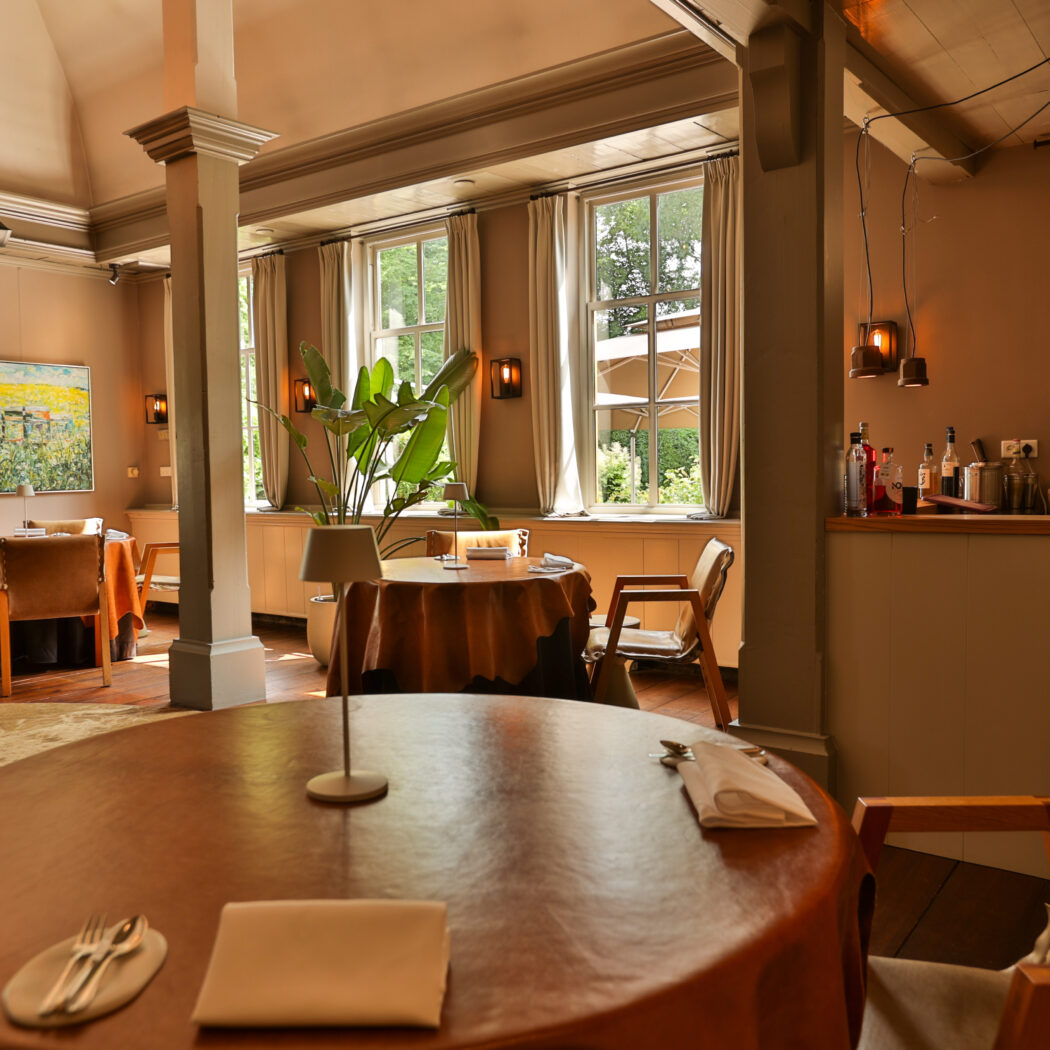 Warm verlicht restaurant met ronde, gedekte tafels, grote ramen en een weelderige plant.
