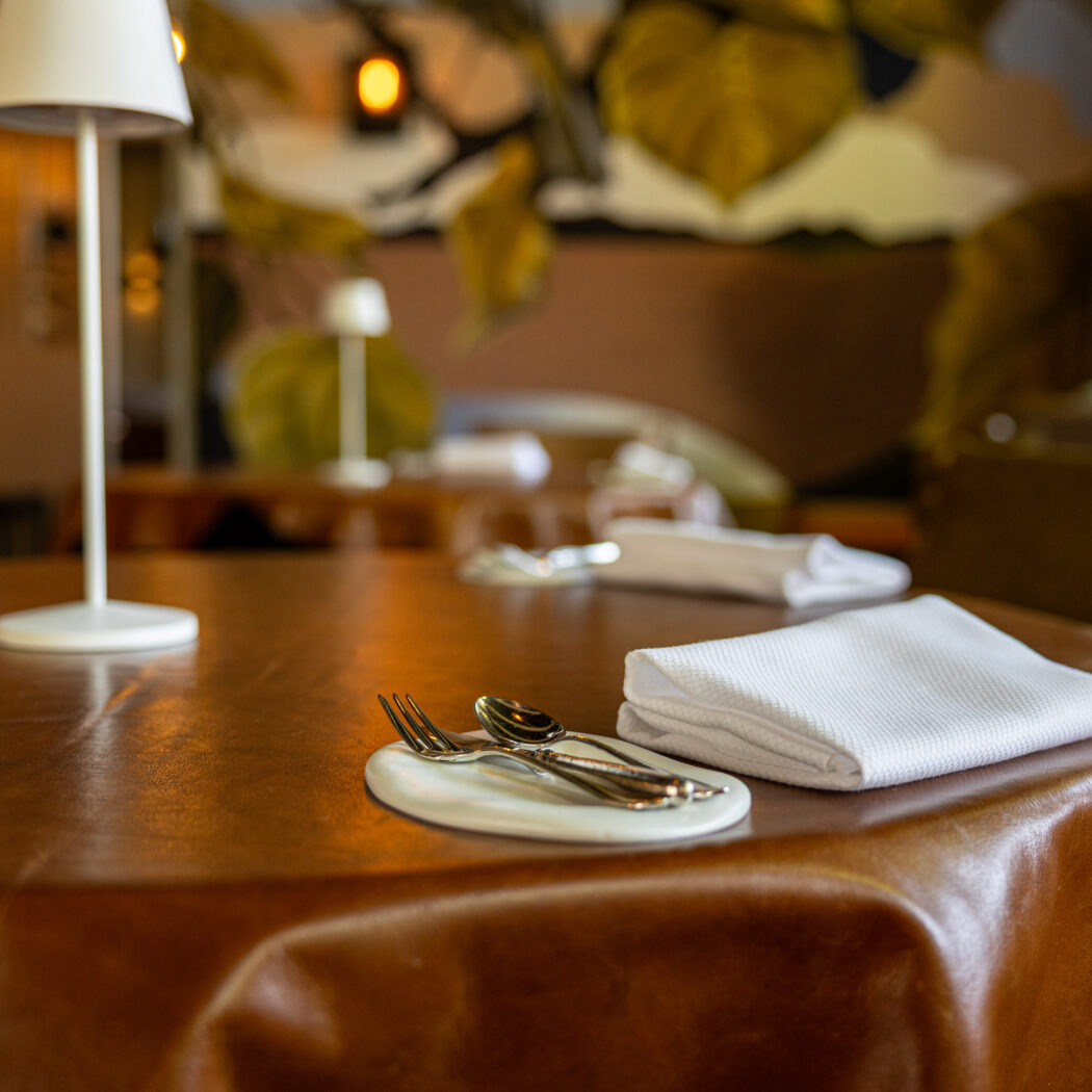 Een gedekte restauranttafel met bruin lederen tafelkleed, bestek op een wit schaaltje, een witte servet en lamp.