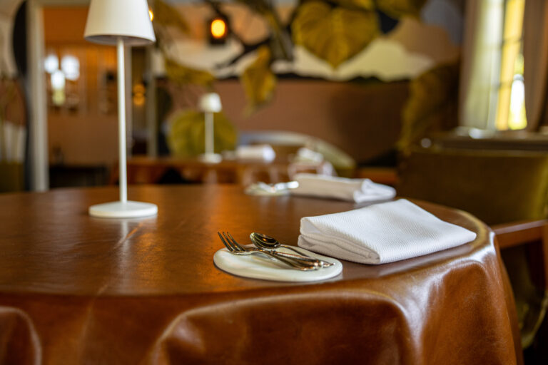 Een gedekte restauranttafel met bruin lederen tafelkleed, bestek op een wit schaaltje, een witte servet en lamp.