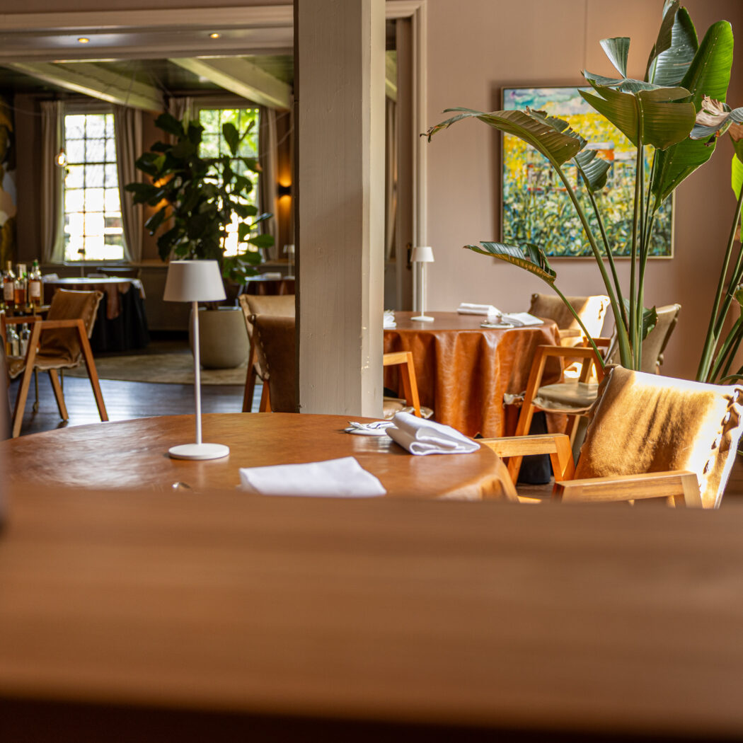 Sfeervol restaurantinterieur met houten tafels, oranje tafelkleden, planten en stoelen met vacht.