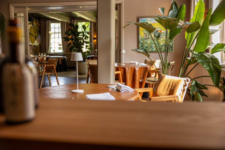 Sfeervol restaurantinterieur met houten tafels, oranje tafelkleden, planten en stoelen met vacht.