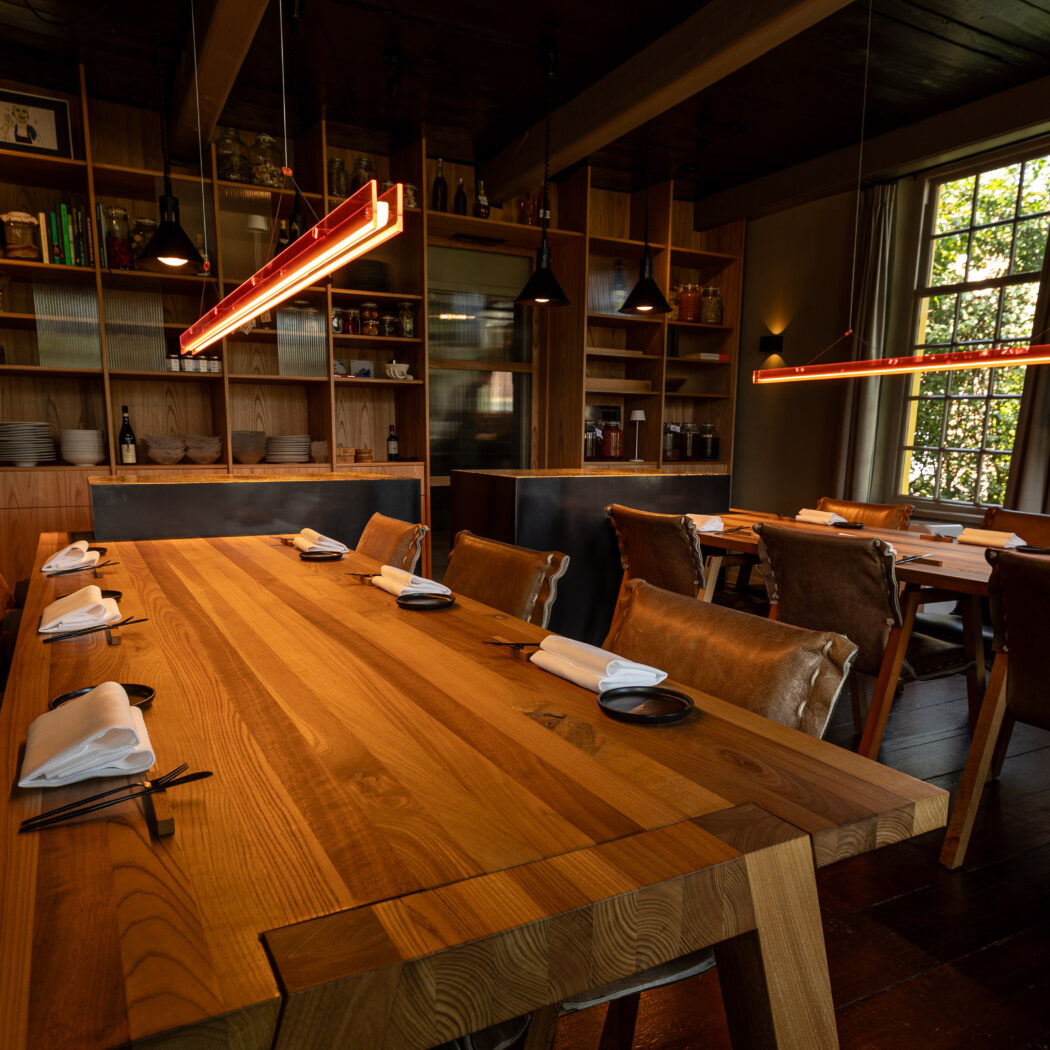 Sfeervolle eetzaal met lange eikenhouten tafels en leren stoelen, verlicht door moderne rode hanglampen.