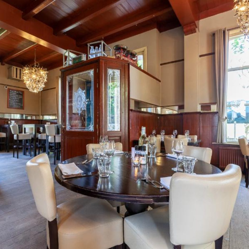 Warm restaurant interieur met donkerhouten balkenplafond, gedekte ronde tafel, beige stoelen en kroonluchters.
