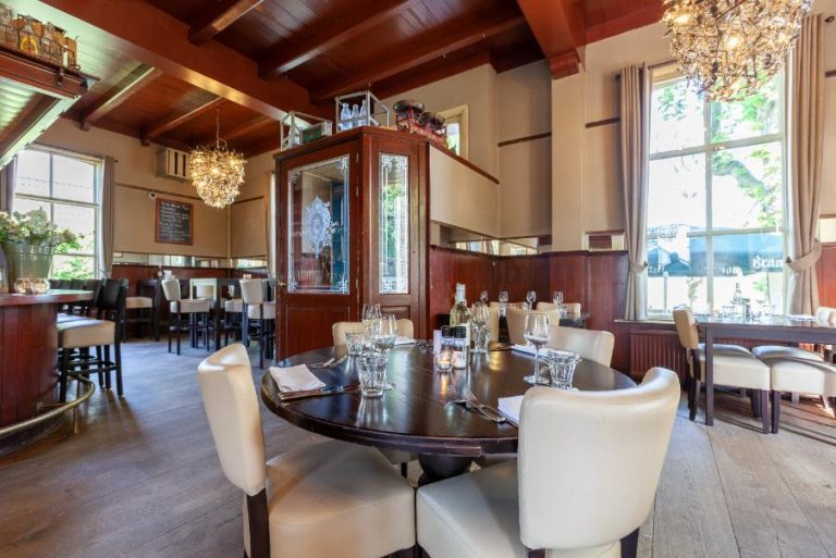 Warm restaurant interieur met donkerhouten balkenplafond, gedekte ronde tafel, beige stoelen en kroonluchters.
