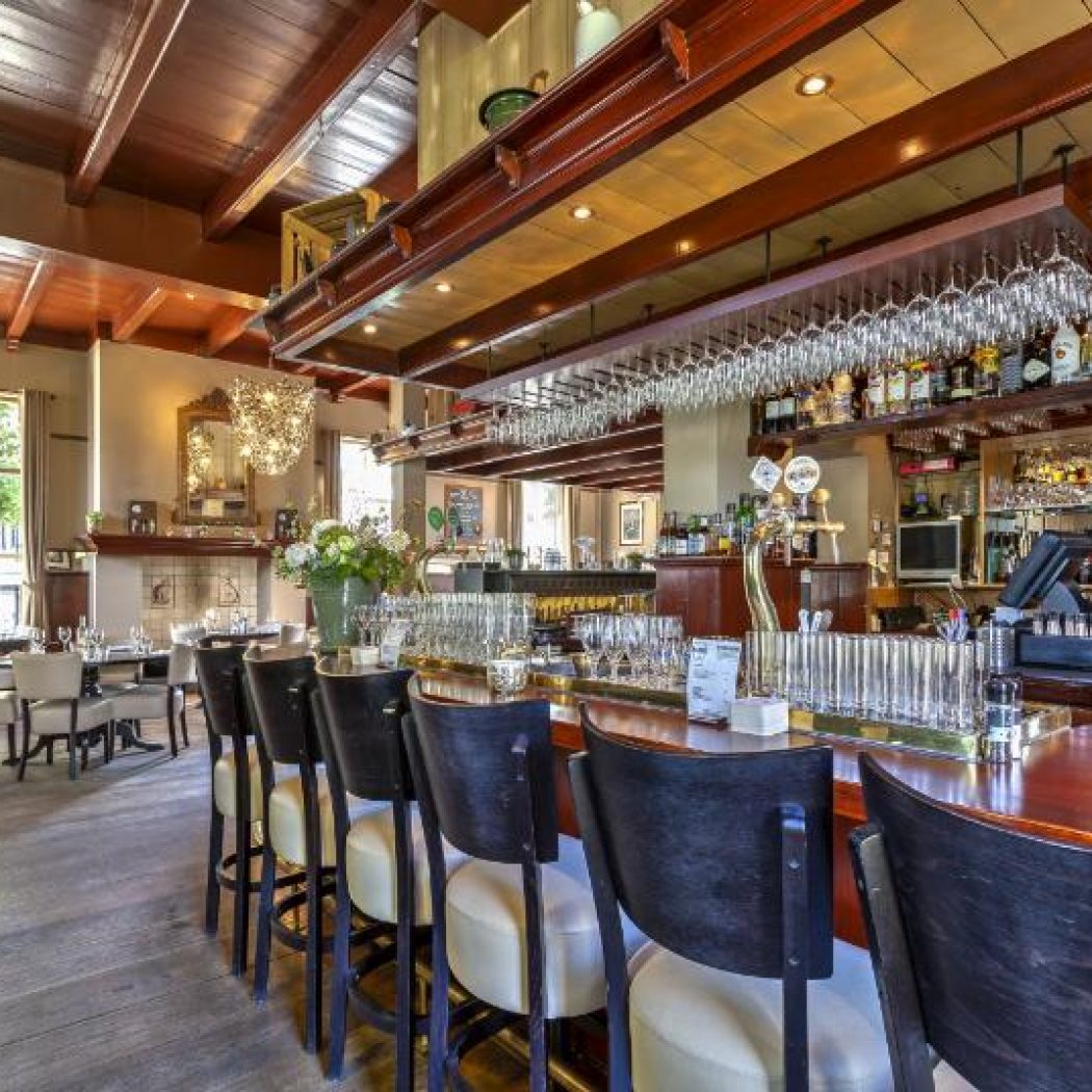 Sfeervolle restaurantbar met donkerhouten toonbank, lichte barstoelen, hangend glaswerk en een groen Brand Lentebock bord.