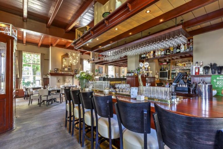 Sfeervolle restaurantbar met donkerhouten toonbank, lichte barstoelen, hangend glaswerk en een groen Brand Lentebock bord.