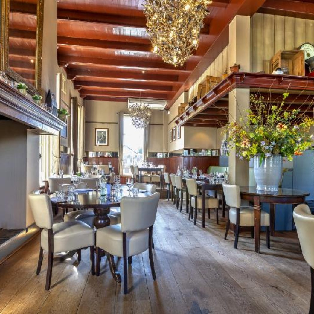 Sfeervol restaurant met houten balkenplafond, kroonluchters, gedekte tafels en een open haard.