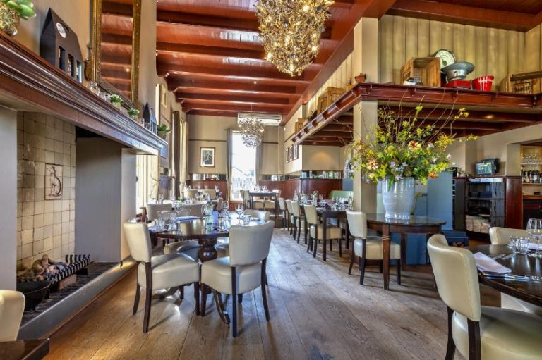 Sfeervol restaurant met houten balkenplafond, kroonluchters, gedekte tafels en een open haard.