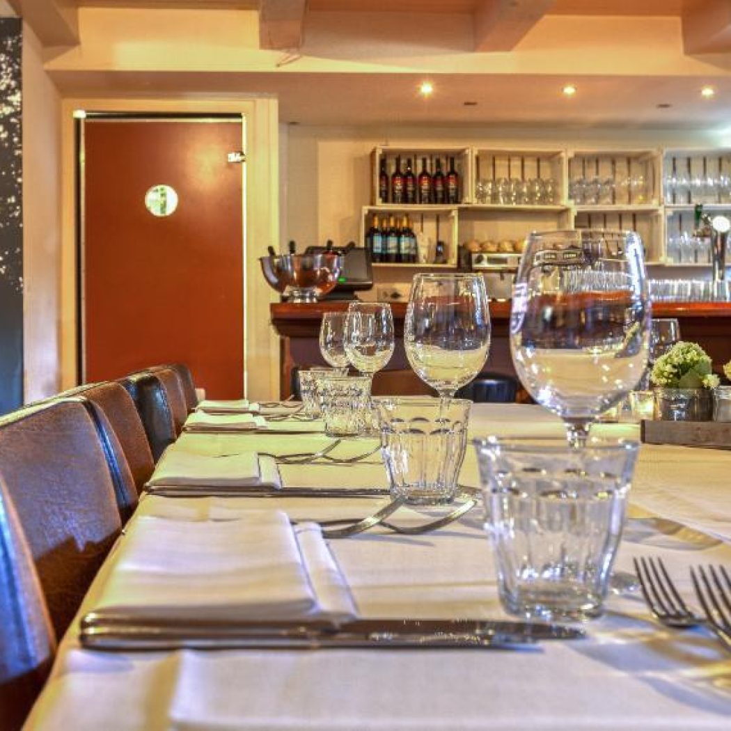 Een lange gedekte restauranttafel met glazen en bestek, bruine leren banken en een bar op de achtergrond.