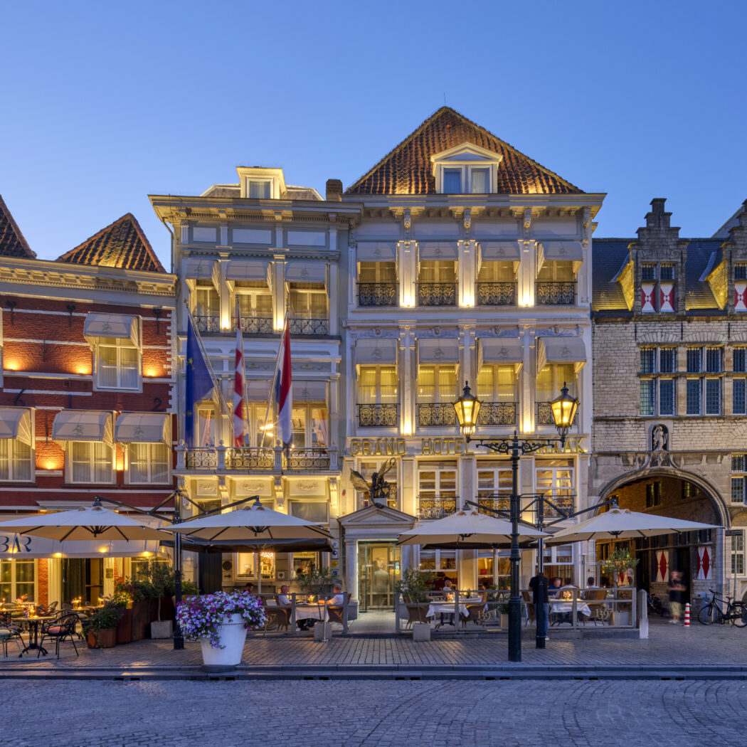 Brano Hotel en historische panden met verlichte terrassen en vlaggen aan geplaveide straat bij schemering.
