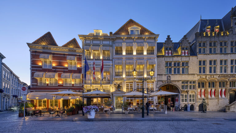 Brano Hotel en historische panden met verlichte terrassen en vlaggen aan geplaveide straat bij schemering.