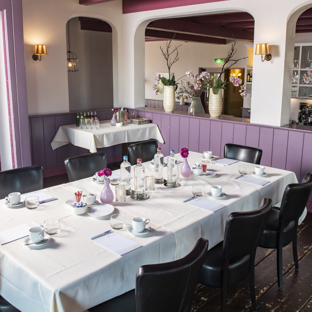 Lange vergadertafel met witte tafelkleden en zwarte stoelen in een paarse zaal, klaar voor een bijeenkomst.