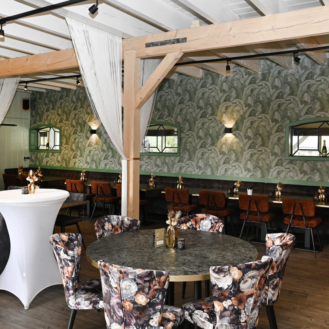 Sfeervol horeca interieur met houten balken, witte doeken, groen bladprint behang, fluwelen banken, ronde tafels en statafels.