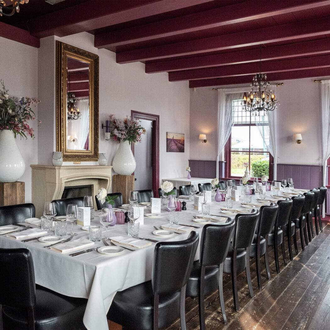 Lange, gedekte tafel met zwarte stoelen in een elegante eetzaal met donkerrode balken en een spiegel boven een schouw.