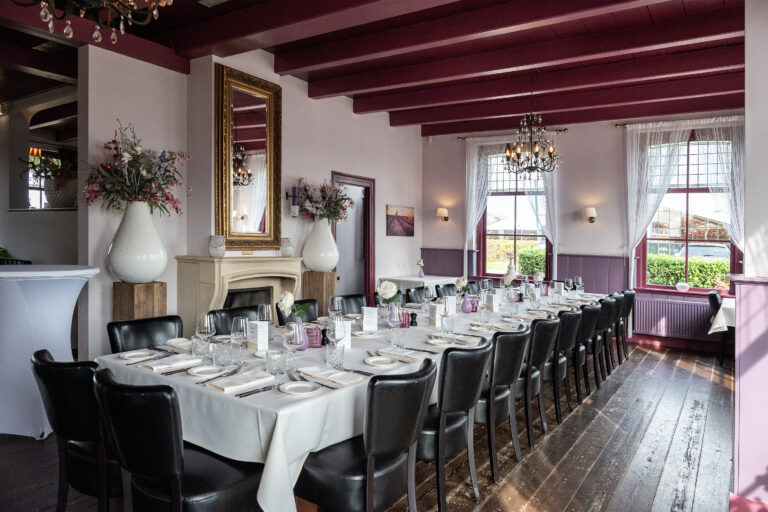 Lange, gedekte tafel met zwarte stoelen in een elegante eetzaal met donkerrode balken en een spiegel boven een schouw.