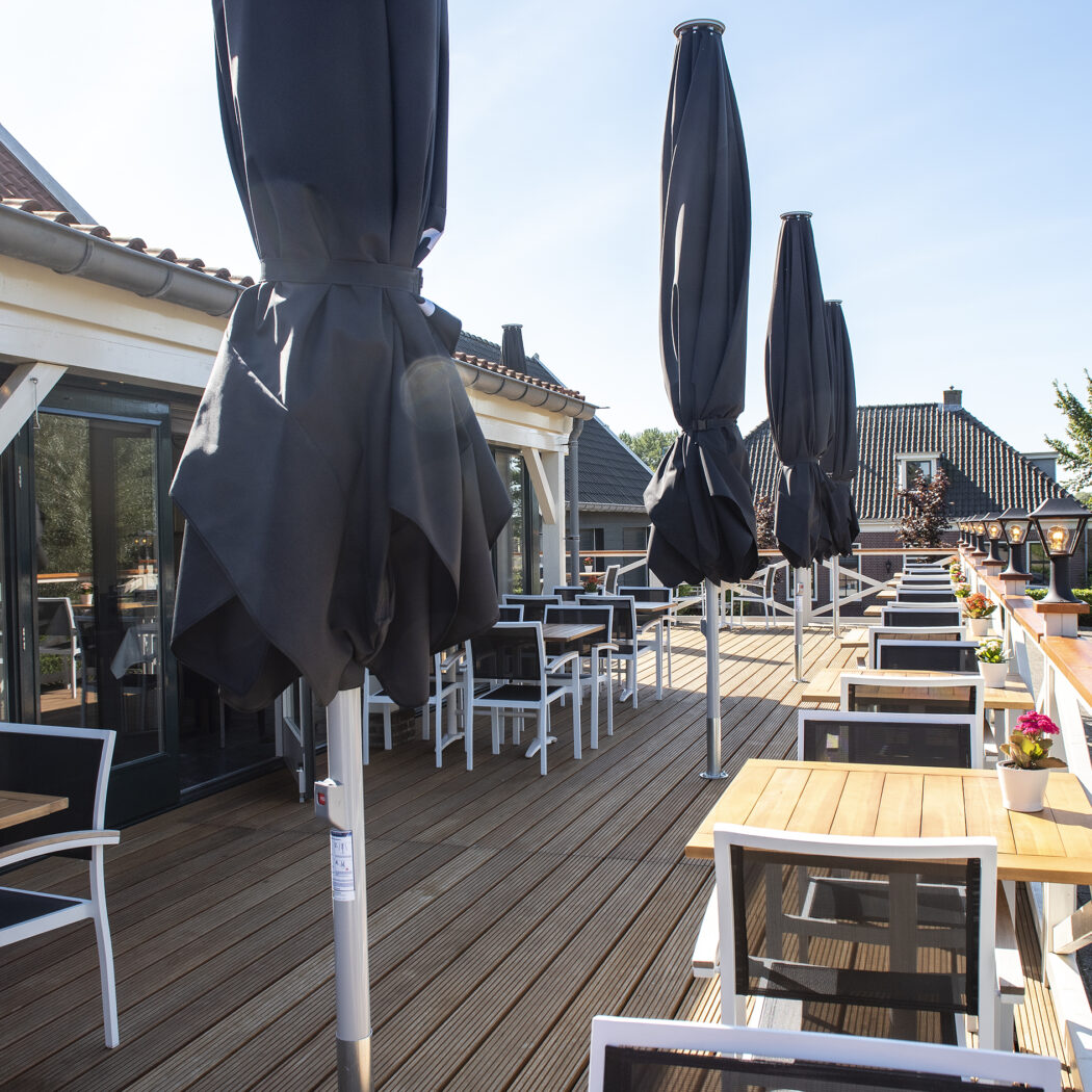 Zonnig houten terras met rijen tafels, stoelen, gesloten zwarte parasols en lantaarns.