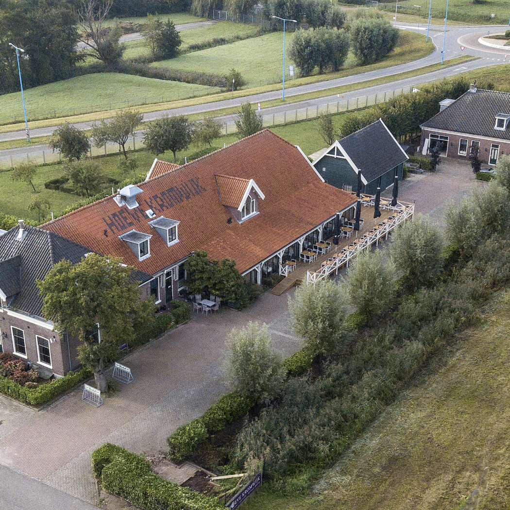 Hoeve Kroonwijk, een gebouw met rood dak en buitenterras, omgeven door groene velden en een weg.