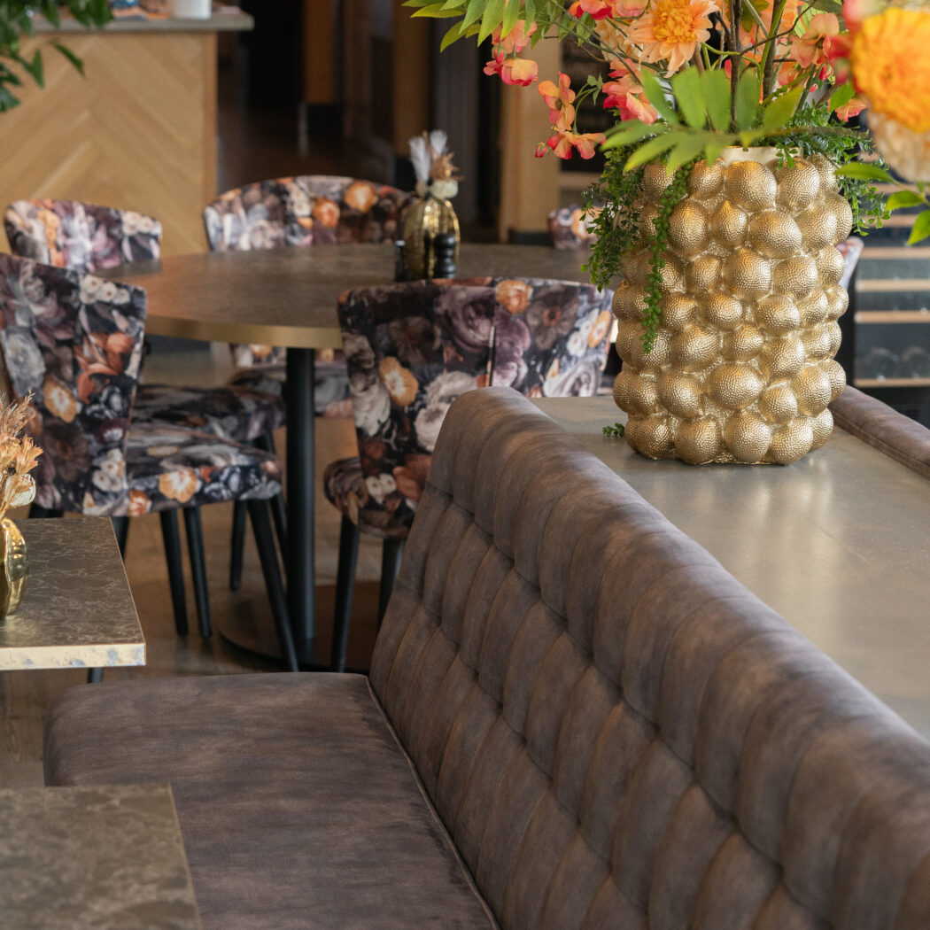 Sfeervol restaurantinterieur met fluwelen loungebank, ronde tafels, stoelen met bloemdessin en gouden vaas met oranje bloemen.
