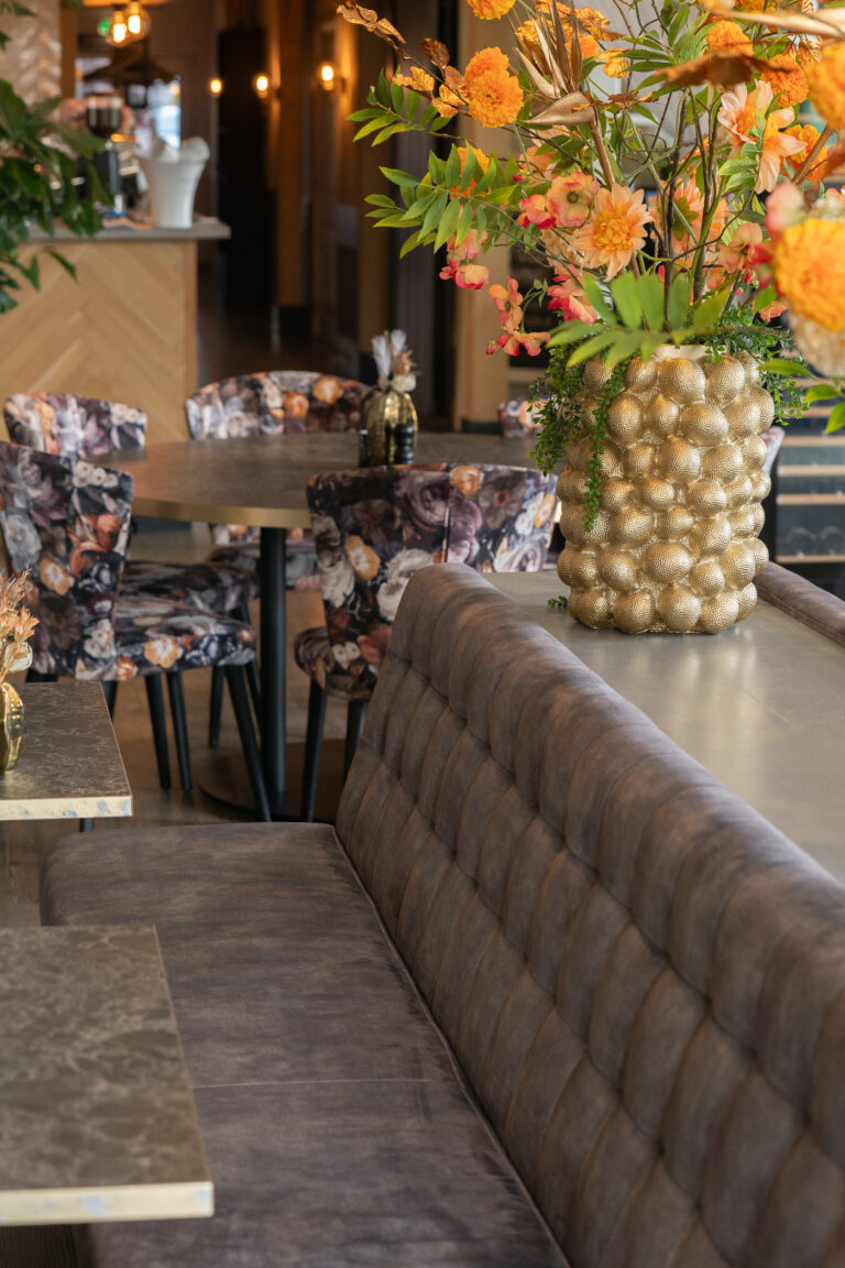 Sfeervol restaurantinterieur met fluwelen loungebank, ronde tafels, stoelen met bloemdessin en gouden vaas met oranje bloemen.