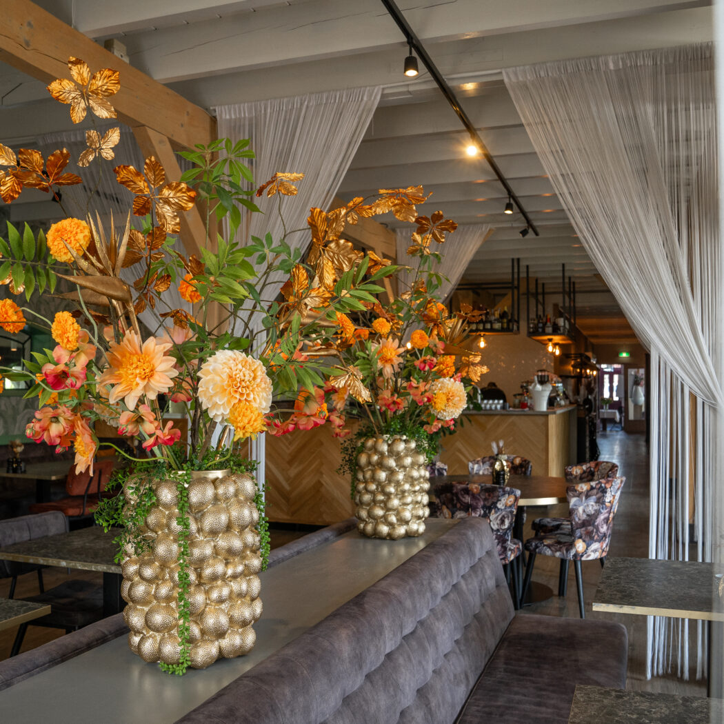Grote bloemstukken met oranje en gouden bloemen in gouden bubbelvazen op een fluwelen bank in een stijlvol restaurant.