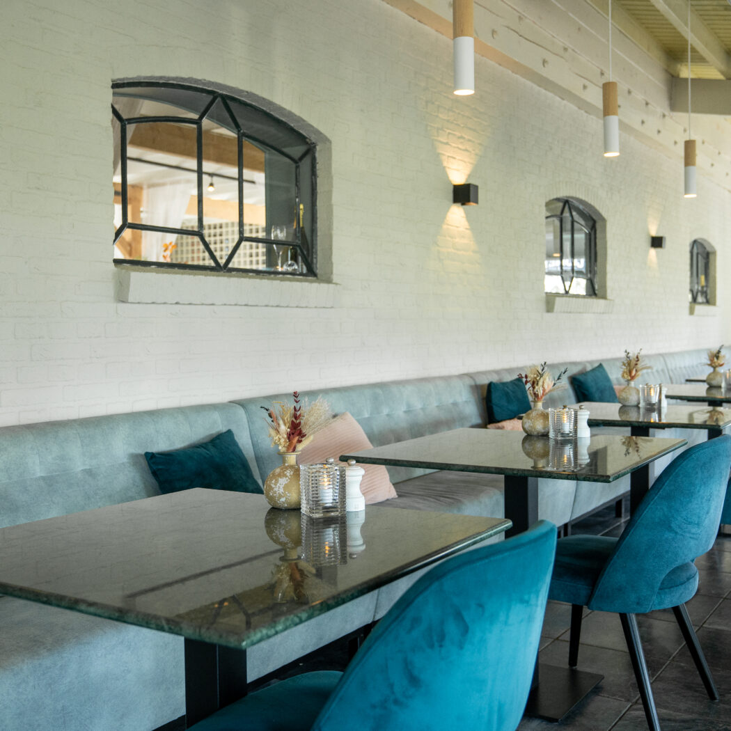 Stijlvol restaurantinterieur met lange lichtblauwe bank, donkere marmeren tafels en teal fluwelen stoelen.