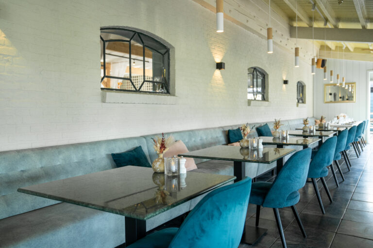 Stijlvol restaurantinterieur met lange lichtblauwe bank, donkere marmeren tafels en teal fluwelen stoelen.