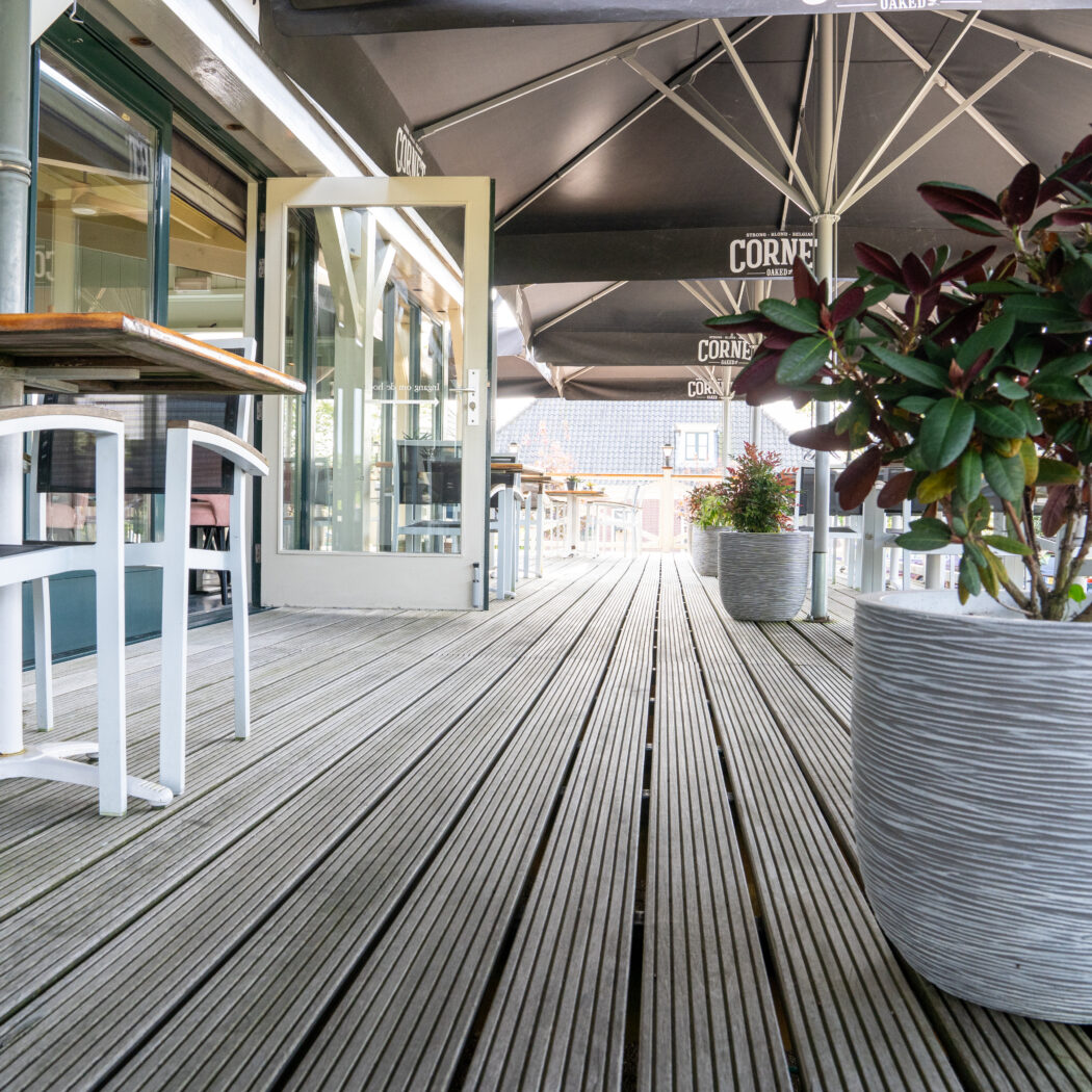 Ruim houten terras met witte tafels en stoelen, grote planten en zwarte parasols van Corne Bakkers.