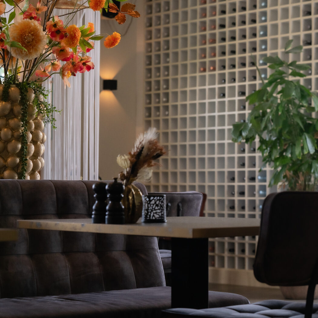 Sfeervol restaurant interieur met een gouden vaas met oranje bloemen, donkere zitbanken en wijnrekken.