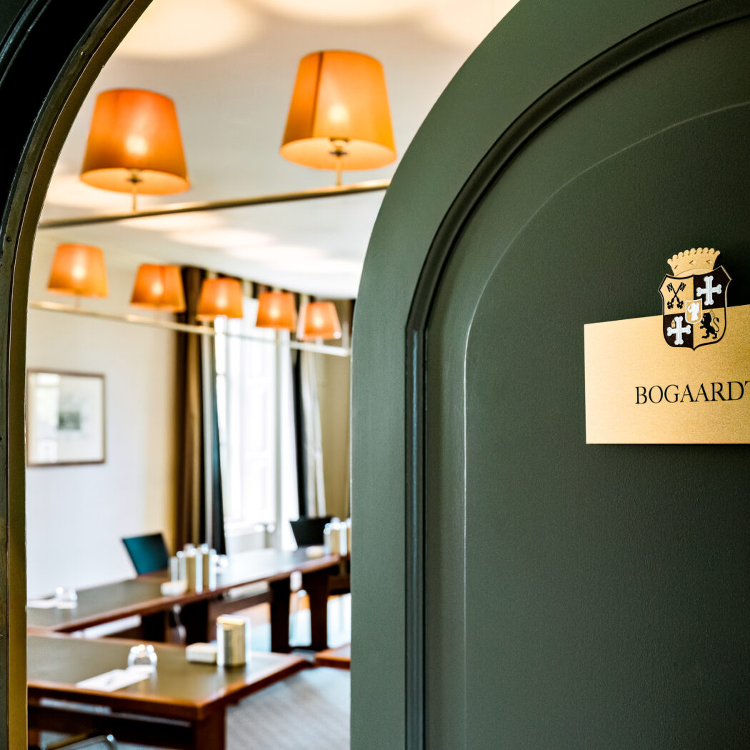 Donkergroene deur met gouden BOGAARDT plaque en wapen, onthult een vergaderzaal met tafels en oranje lampen.