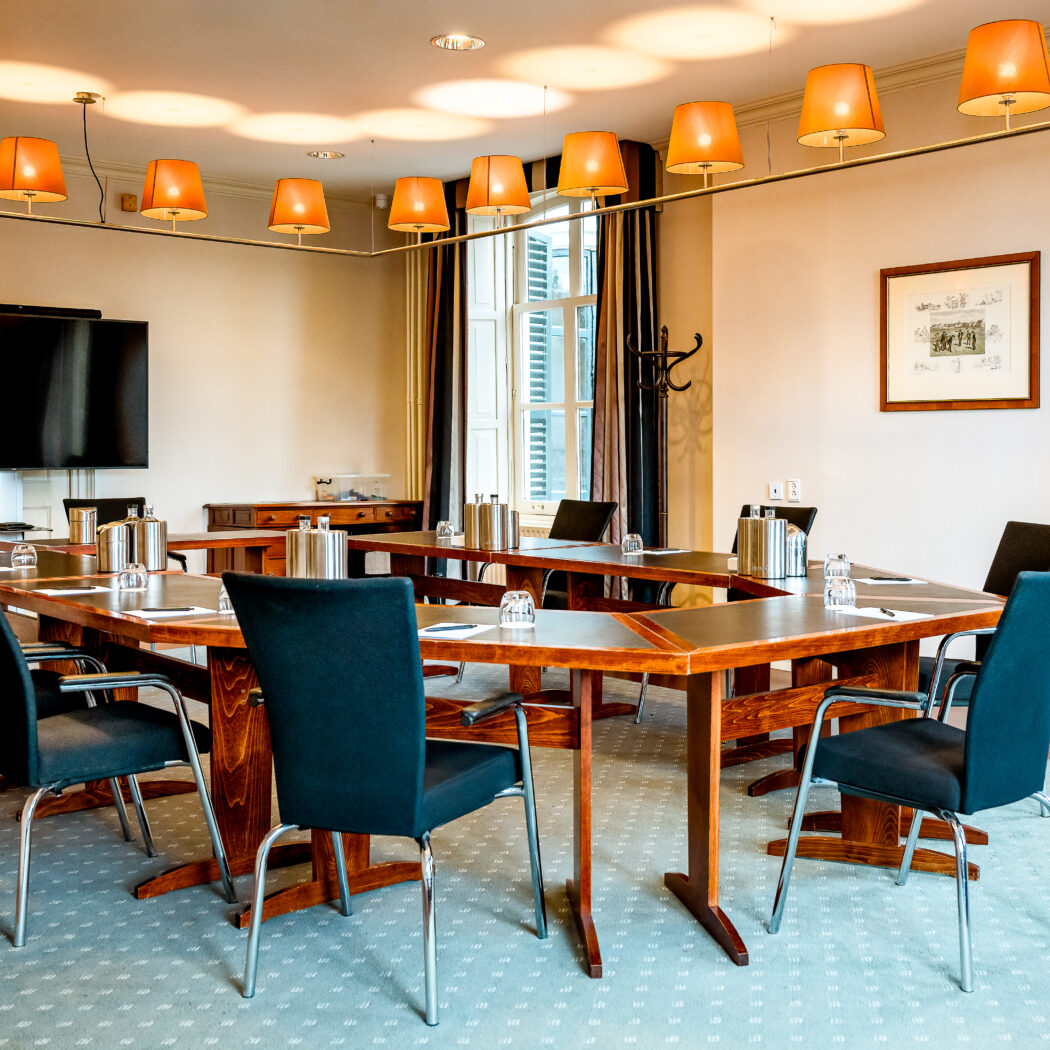 Elegante vergaderzaal met U-vormige houten vergadertafel, zwarte stoelen, tv en oranje lampen.