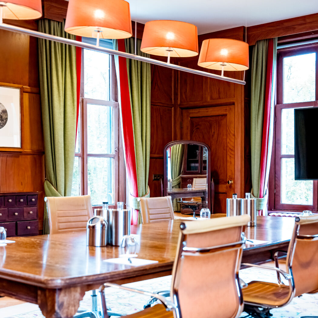 Een klassieke vergaderzaal met lange houten tafel, lichtbruine stoelen, houten lambrisering, oranje lampen en tv.