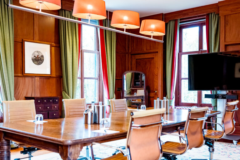 Een klassieke vergaderzaal met lange houten tafel, lichtbruine stoelen, houten lambrisering, oranje lampen en tv.