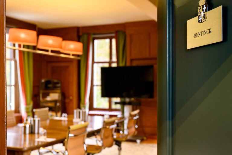 Gouden naambord BENTINCK met een wapen aan een donkergroene muur, met uitzicht op een vergaderzaal met houten tafel en tv.